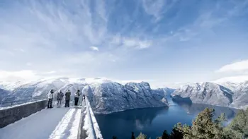 A group stare over a view point in Myrkdalen, looking at a vast landscape of snowy mountains, lush fields and wide, flowing rivers all under a bright blue sky.